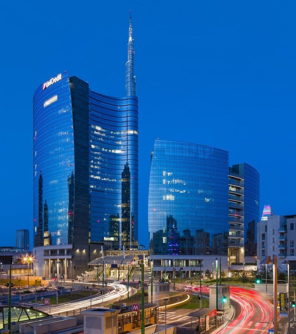 UniCredit: Glass towers with a tall spire against a deep blue evening sky, traffic light trails below