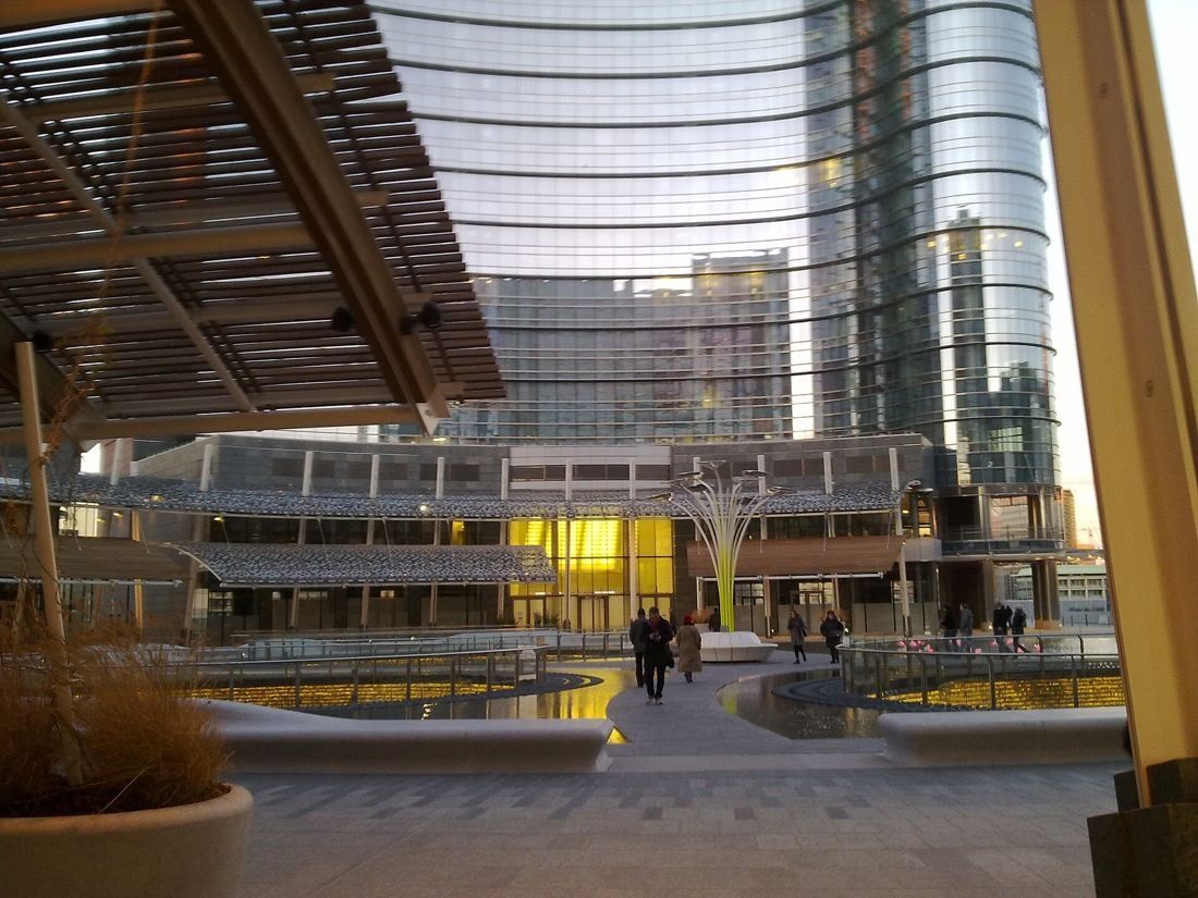 UniCredit: Round plaza with pools and lit shopfronts under a curved glass tower
