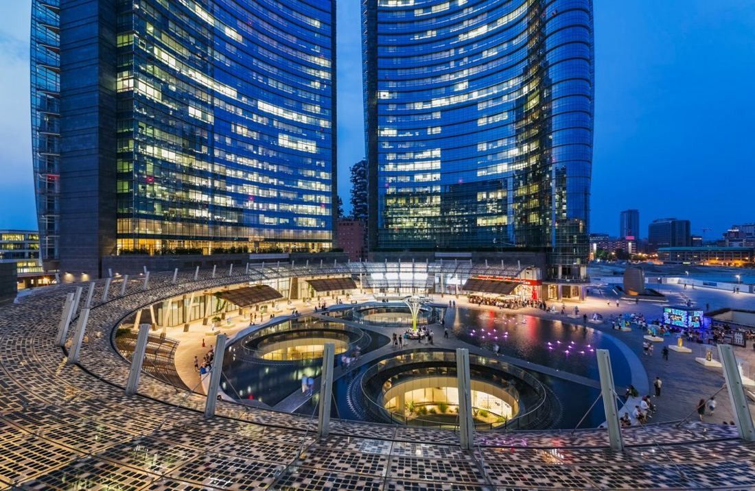 Two curved glass towers lit at dusk above a circular sunken courtyard with glowing openings