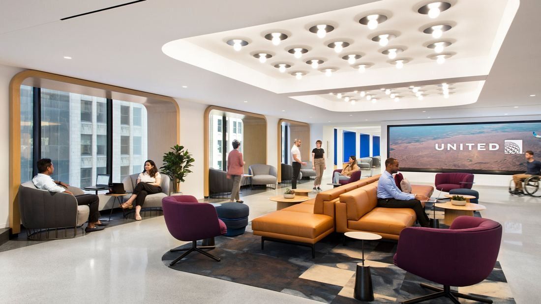 United Airlines: Bright lounge with orange and purple sofas, a lit ceiling and a wide screen at the end