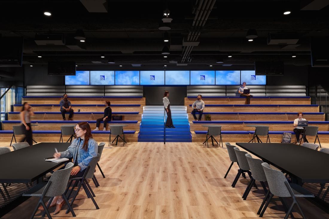 United Airlines: Large room with wooden bleacher seating, a row of screens above and tables on a wood floor