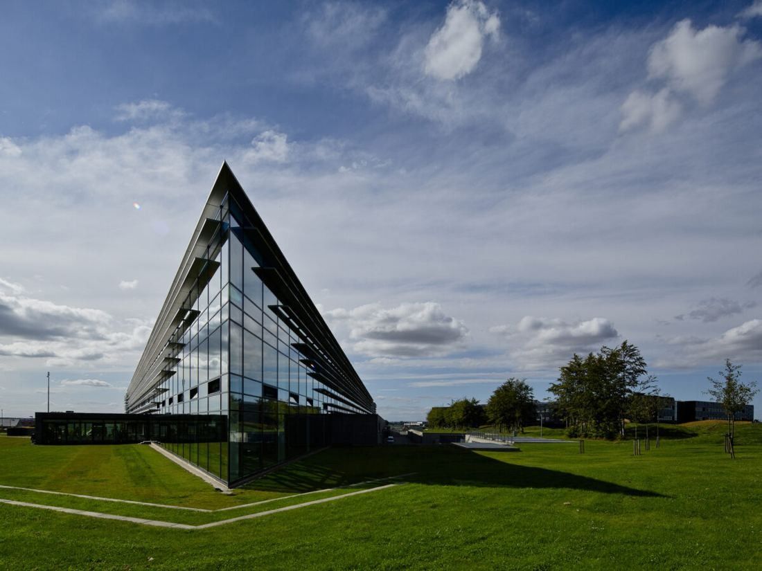 A dark glass building ending in a sharp triangular point above a green lawn under a cloudy sky