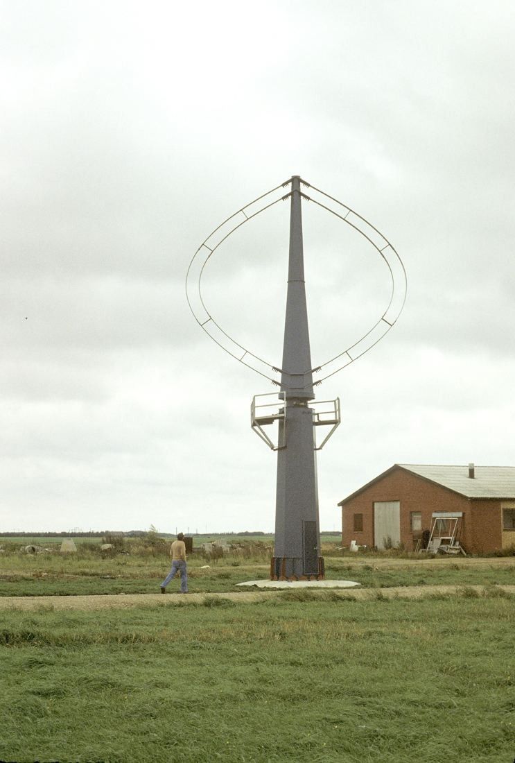 Slender vertical-axis wind turbine with two curved blades standing in a field near farm buildings