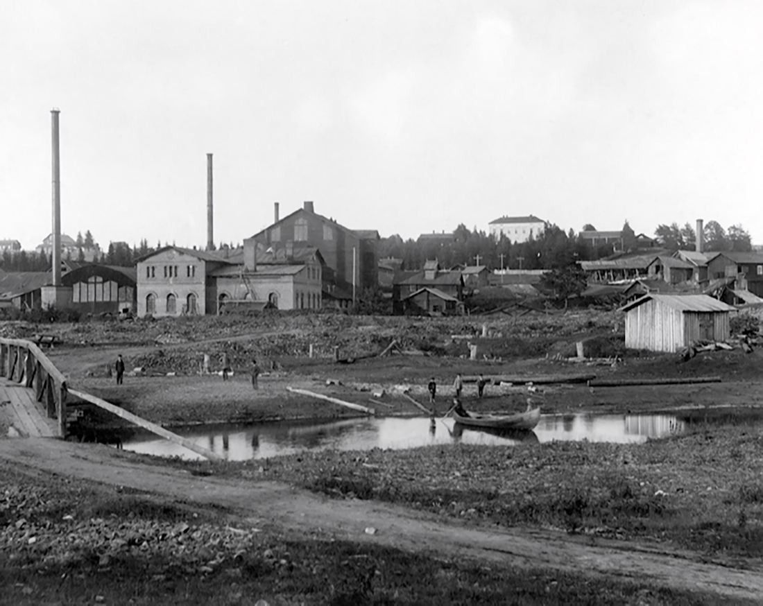 Black-and-white photo of low wooden mill buildings and tall chimneys beside a pond