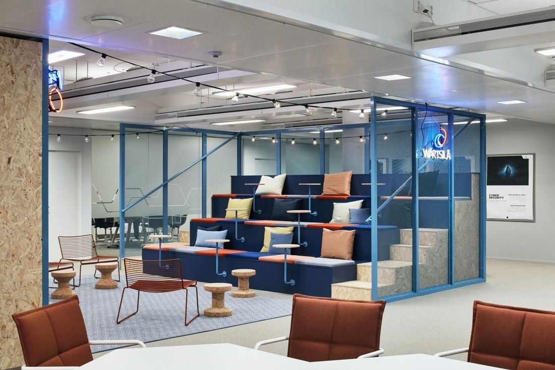 Wärtsilä: Blue-framed glass box with tiered seating and cushions in an open office, red chairs in the foreground