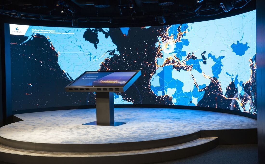 Wärtsilä: Dark room with a curved wall-size screen showing a world map and a touchscreen stand on a round platform