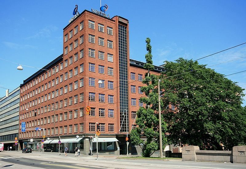 Red-brick office building with a tall glazed stair tower and a Wärtsilä logo on the roof, on a street corner