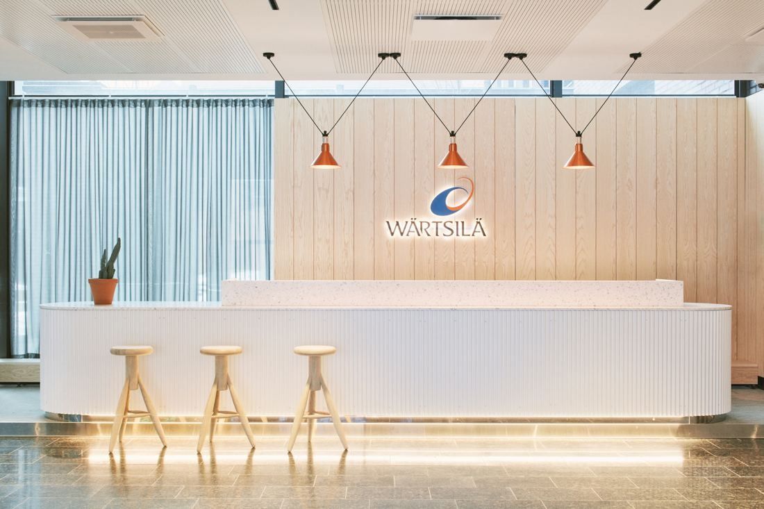 Wärtsilä: White reception desk with bar stools and three copper pendant lamps in front of a pale wooden wall bearing the Wärtsilä logo
