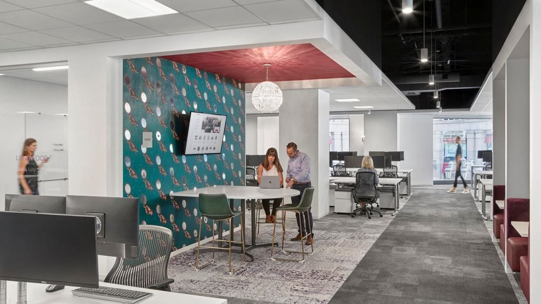 Wayfair: Huddle area with a round table under a patterned wall, next to rows of desks with monitors
