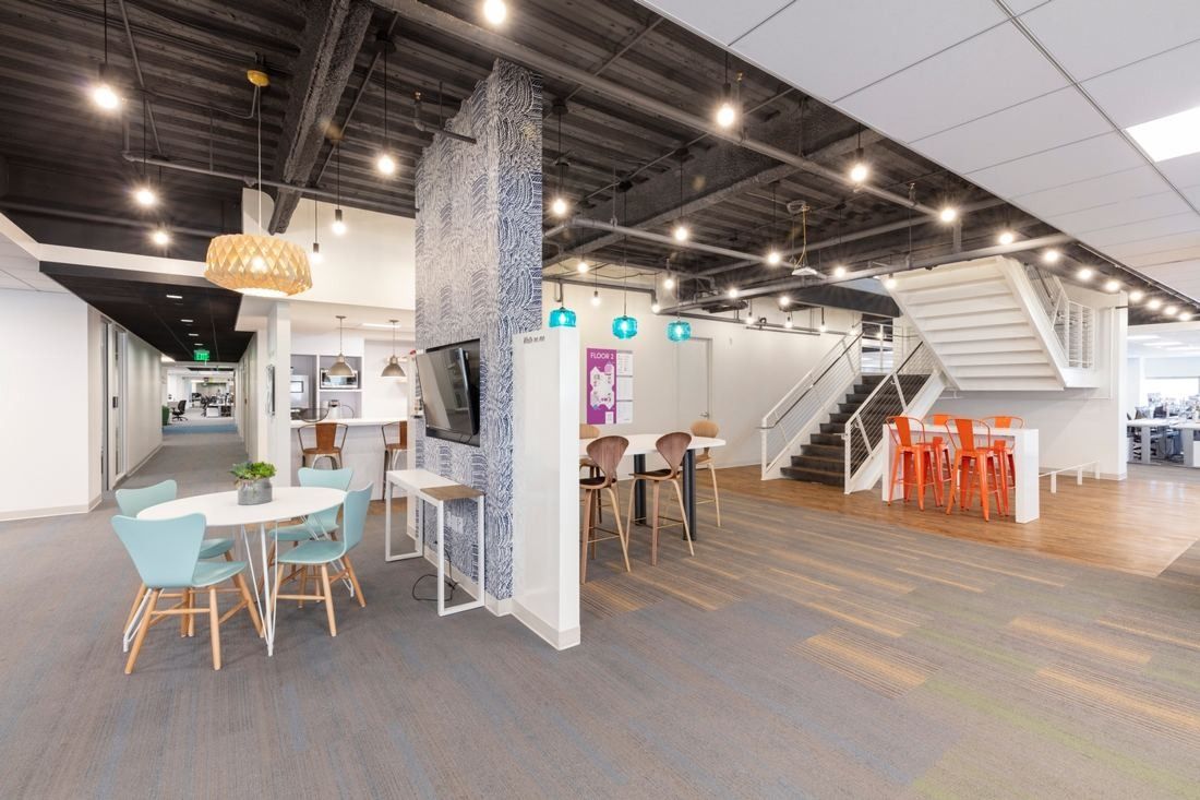 Wayfair: Open office floor with café tables, a tiled column and a white stair rising at the back