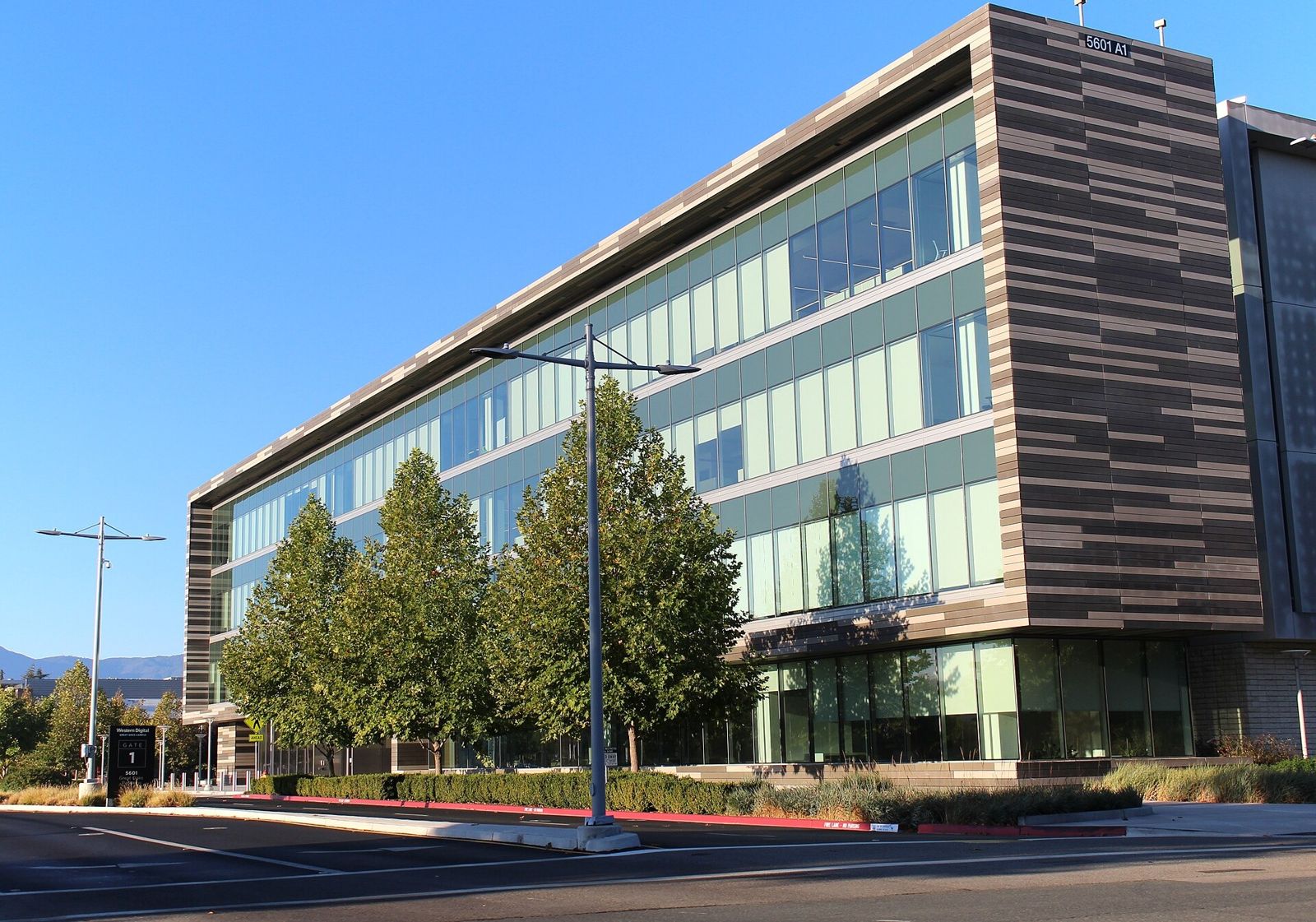 A four-storey glass office building with a striped wood-tone end wall behind young trees