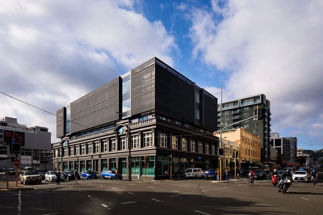Corner view of a two-storey heritage brick facade with a dark multi-storey glass and mesh block rising above it, under a blue sky