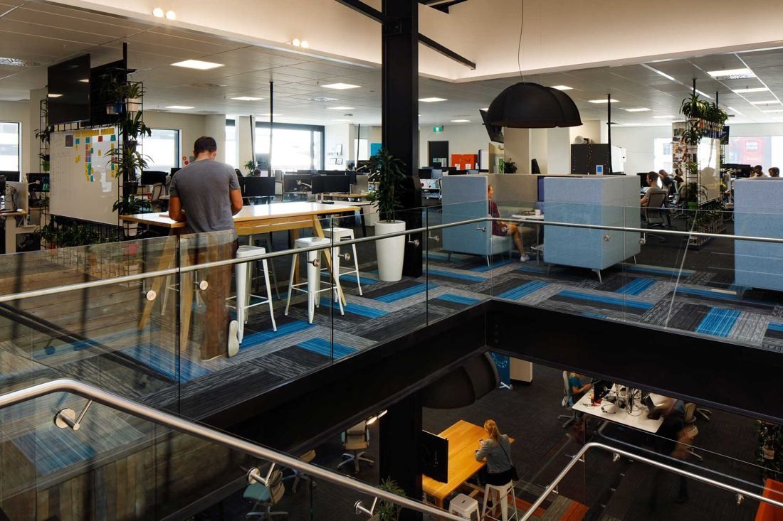 Xero: Open floor seen from the stair balustrade with high tables, booths and desks on the level below