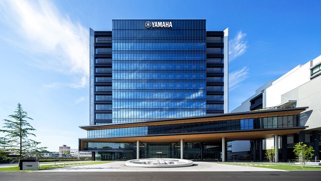 A tall glass office tower with a low, wide wing in front and a landscaped plaza under a blue sky