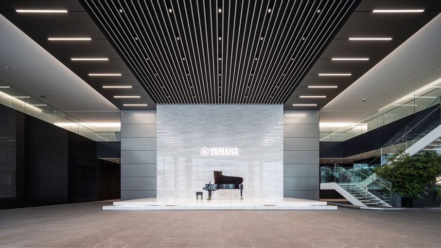Yamaha: A tall, open entrance hall with a slatted ceiling and a grand piano in front of a white wall