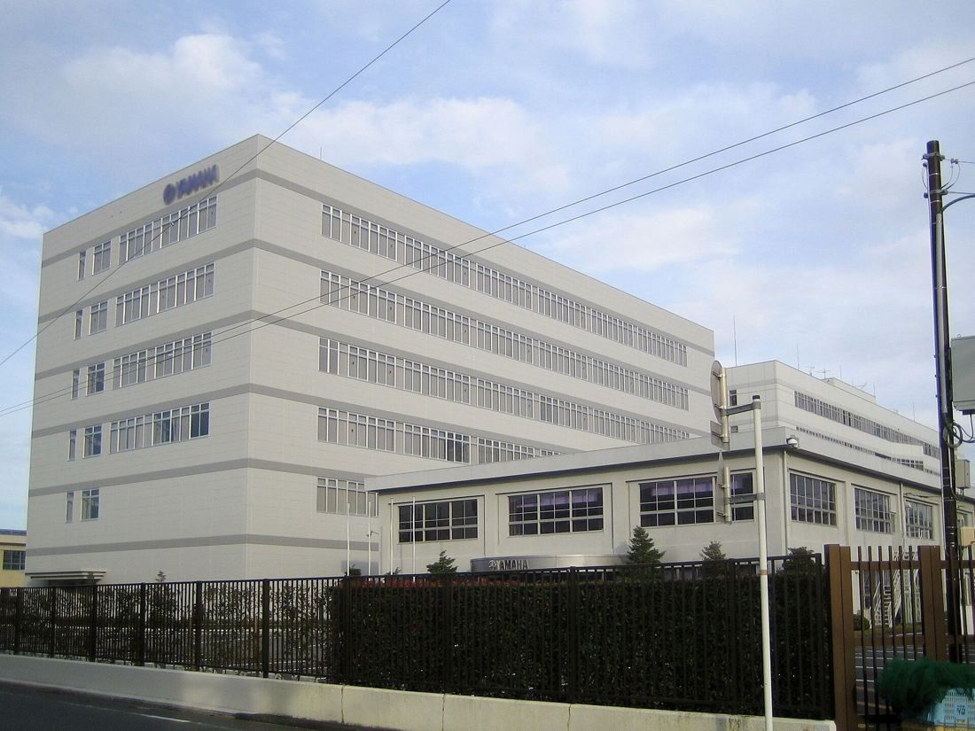 Yamaha: A white multi-story building behind a fence and an older low building at a street corner