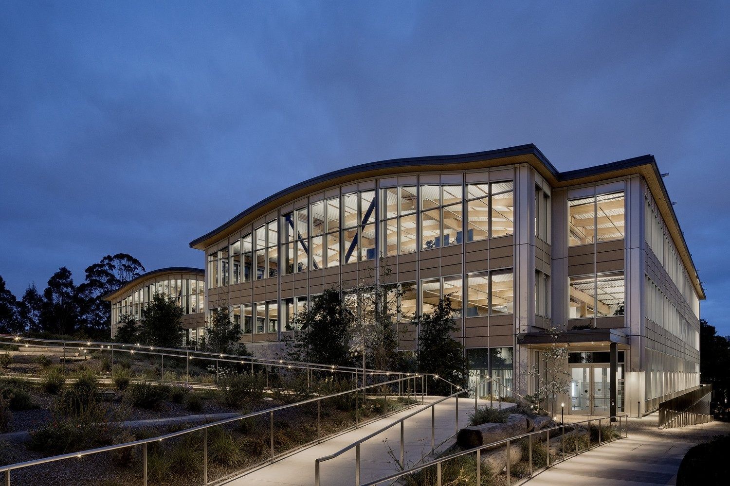 YouTube: Glass-fronted building with a curved roof lit at dusk above a landscaped path