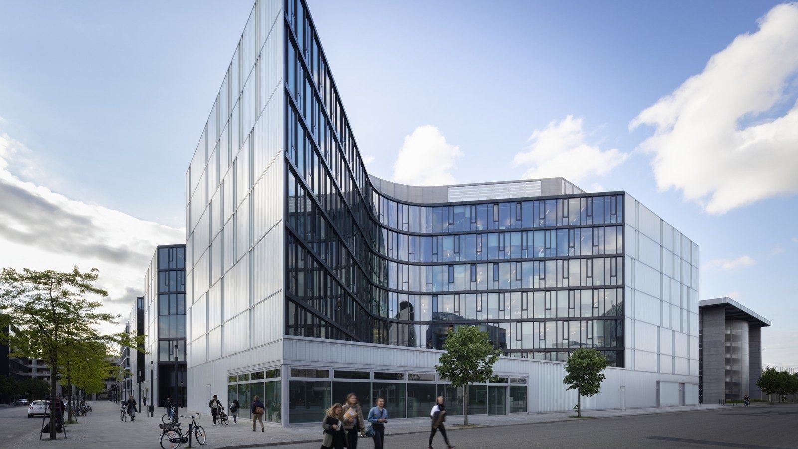 Corner of a glass office building with a dark curved facade and a silver panelled wing under a blue sky