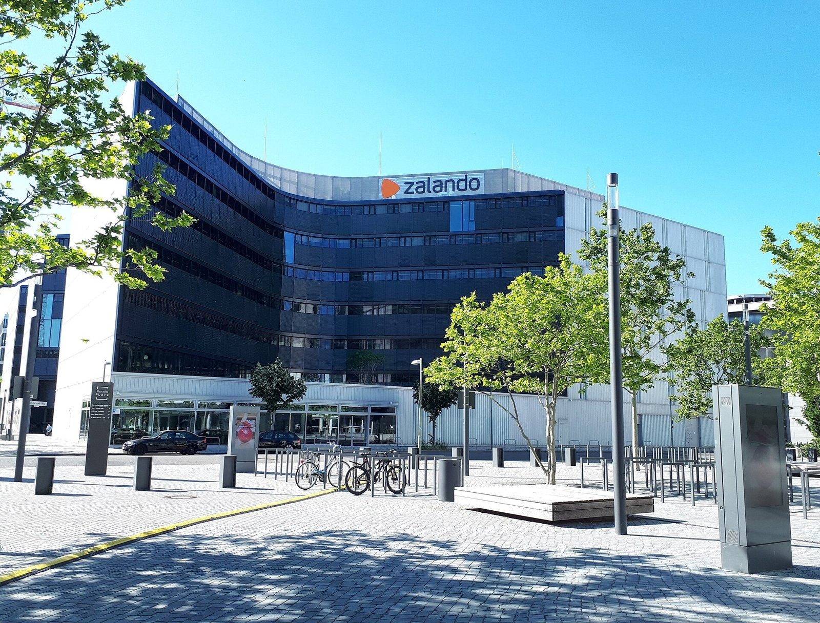 Zalando: Curved dark glass facade with the Zalando sign behind a paved plaza with young trees