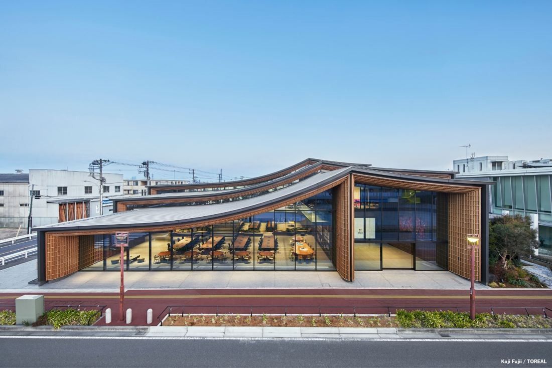 Low building with a wavy layered roof and glass front facing a street under a blue sky