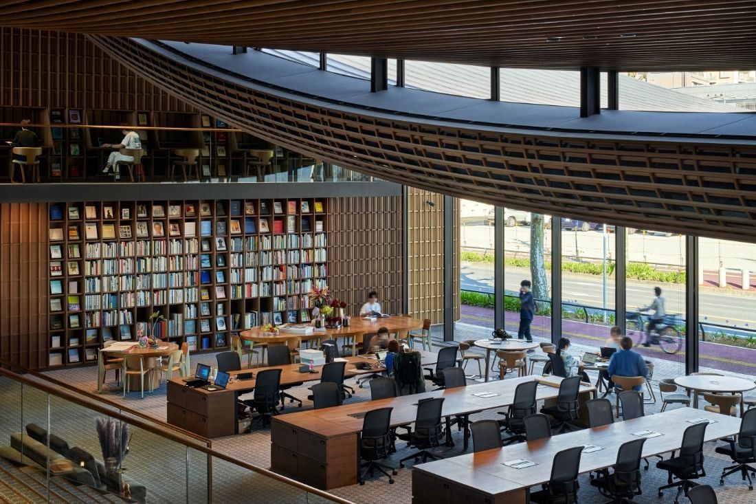 ZOZO: Workspace with long desks, a tall bookshelf wall and a mezzanine under a curved wooden ceiling, street visible through glass