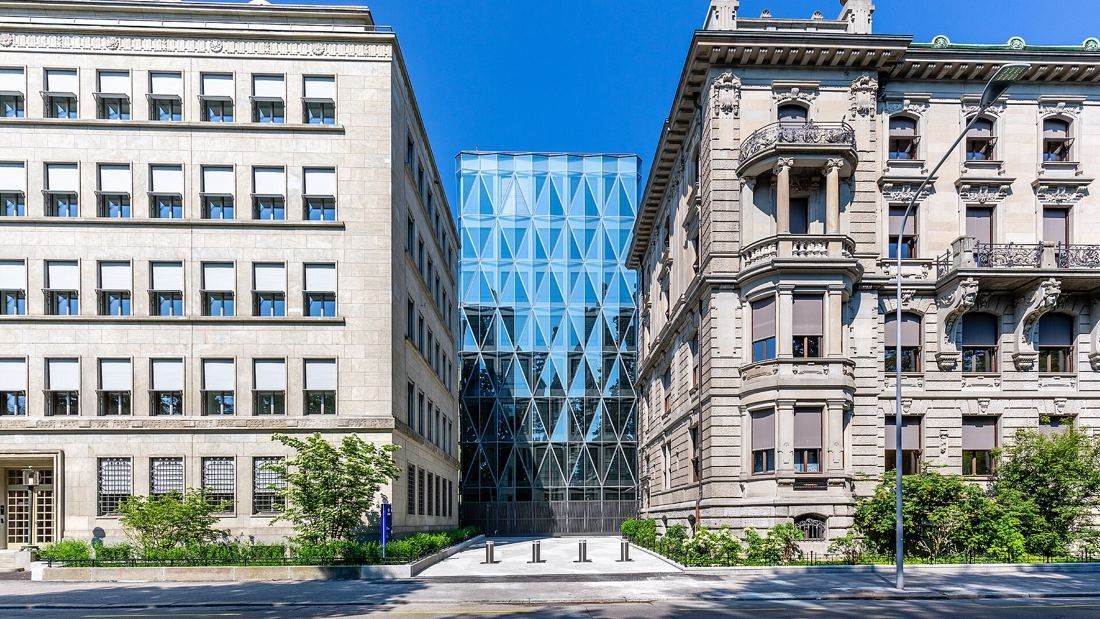 A faceted blue glass building set between a pale stone office block and an ornate historic facade, seen from the street