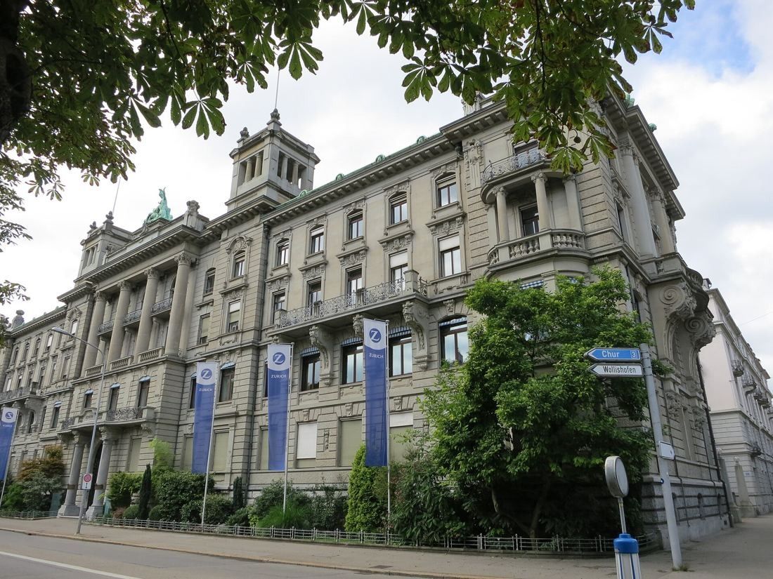 Zurich Insurance Group: Large historic stone building with towers on a street corner, blue flags along the front and trees